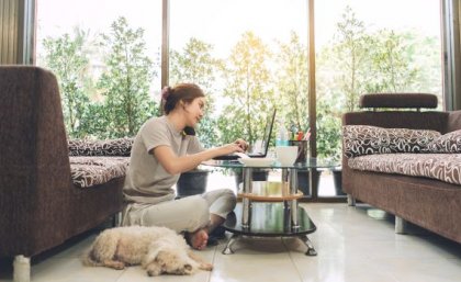 A woman sits on the floor in front of a low table using a phone and laptop computer, a dog is next to her.